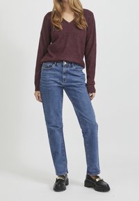 Pull col en V marron en matière douce, associé à un jean bleu en denim taille haute. Mocassins à plateforme noirs avec des accents dorés.