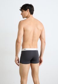 Graue Boxershorts mit einem weißen Bund, der schwarzen "BOSS"-Text zeigt, auf einem männlichen Modell von hinten dargestellt. Glatte Stoffstruktur.