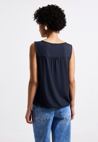 Marinblå ärmlös topp med en samlad ryggdetalj, tillverkad av ett slätt tyg. Kombineras med ljusblå jeans som har bakfickor och en avslappnad passform.