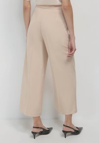 Pantalons larges beiges en tissu lisse, présentant un design épuré avec des coutures latérales et un ourlet droit. Des chaussures à talons noires sont portées.