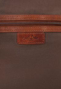 Bruin stoffen oppervlak met een horizontale ritssluiting en een genaaide leren patch-label onder de ritssluiting.
