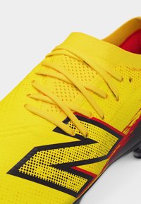 Κίτρινο αθλητικό παπούτσι με υφασμένο επάνω μέρος, κίτρινες κορδόνια και μεγάλο μαύρο και κόκκινο στυλιζαρισμένο λογότυπο "N" στο πλάι.