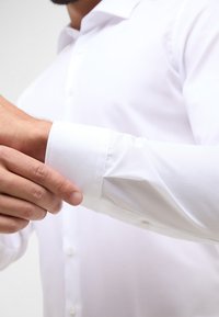 Camicia bianca a maniche lunghe con texture liscia, chiusura con bottoni sul davanti e colletto aderente. I polsini sono semi-aderenti, mostrando la lucentezza del tessuto.