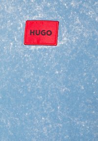 Světle modré bavlněné tričko s oprýskanou texturou, s červeným obdélníkovým nášivkem se slovem "HUGO" v černém písmu.