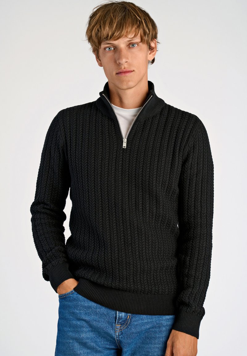Schwarzer Strickpullover mit hohem Kragen und Reißverschluss vorne. Strukturiertes Muster mit gerippten Bündchen und Saum. Kombiniert mit blauen Jeans.