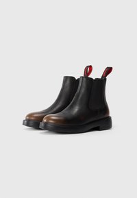 Bottes Chelsea en cuir noir et marron avec des côtés élastiques noirs et des tirettes rouges étiquetées "Camper" sur fond blanc.