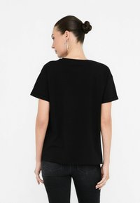 T-shirt nera a maniche corte con vestibilità rilassata, materiale in cotone e orli arrotolati. La vista posteriore mostra un design semplice senza grafiche o loghi visibili.