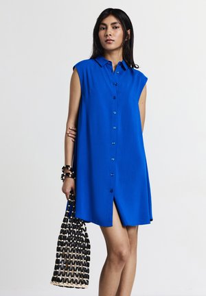 Donna che indossa un vestito senza maniche di un vivace blu con bottoni, mentre tiene in mano una borsa intrecciata nera e beige, su uno sfondo semplice.