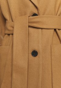 Abrigo de lana color camel con cintura ceñida, que presenta una tela texturizada, dos bolsillos laterales y un cierre de botón negro.