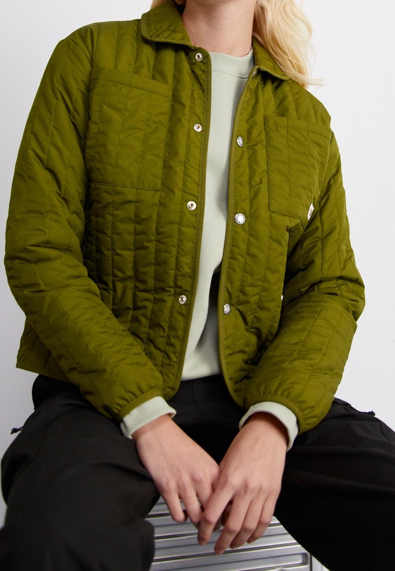 Personne portant une veste matelassée verte avec des boutons-pression, une chemise vert clair en dessous et un pantalon noir, assise les mains reposant sur les genoux.