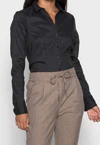 Black button-up shirt in smooth fabric, featuring a collar and long sleeves. Paired with beige plaid trousers with a drawstring.