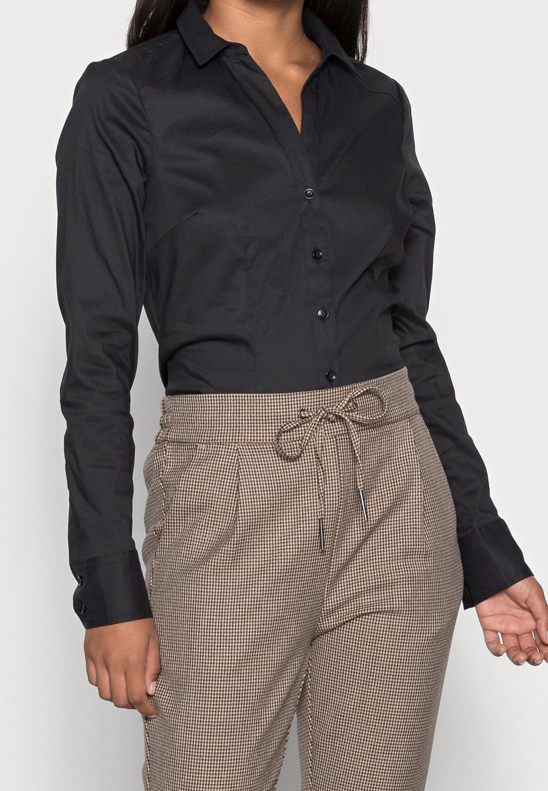 Black button-up shirt in smooth fabric, featuring a collar and long sleeves. Paired with beige plaid trousers with a drawstring.