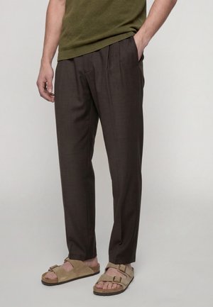 Homme portant un pantalon marron foncé et des sandales Birkenstock beige, une main dans la poche et un pull vert olive.