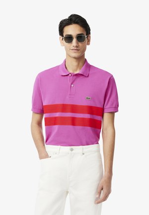 Polo rosa con strisce orizzontali rosse, maniche corte e colletto classico. Indossato con jeans bianchi e occhiali da sole scuri. Presenta un dettaglio con logo.