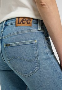 Jean de mezclilla azul claro con un parche de cuero marrón con el logo "Lee" en la parte trasera de la cintura, que cuenta con bolsillos estándar y detalles de costura amarilla.