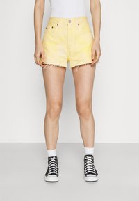 Shorts de mezclilla amarillos con el dobladillo deshilachado, diseño de cinco bolsillos y cierre frontal de botones, combinados con zapatillas negras de caña alta y calcetines blancos.