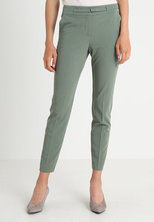 Wąskie zielone spodnie o długości do kostek z paskiem i szlufkami, noszone z jasnoszarymi butami na obcasie z szpiczastym noskiem i białą bluzką.