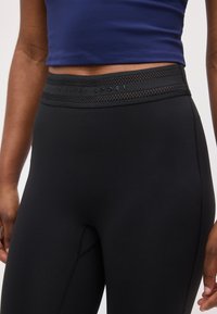 Κοντινό πλάνο ατόμου που φοράει μαύρα κολάν με ψηλή μέση και ζώνη με δίχτυ, και μπλε crop top, με τα χέρια χαλαρά στα πλάγια.