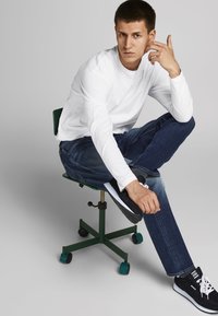 Witte langdikke shirt, blauwe denim jeans, zwarte sneakers. Model zittend op een groene metalen stoel, met één been over het andere gekruist.