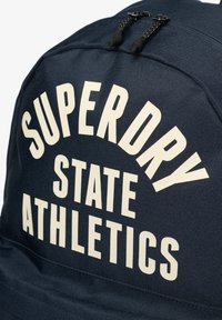 Marineblå stofrygsæk med stor hvid tekst: "SUPERDRY STATE ATHLETICS." Tekstureret overflade med sorte lynlåstræk.