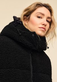 Črna flis jakna z visokim ovratnikom in kapuco z vrvico. Ima sprednji zadrgo, mehko teksturo ter je dobro prilegajoča z modernim dizajnom.