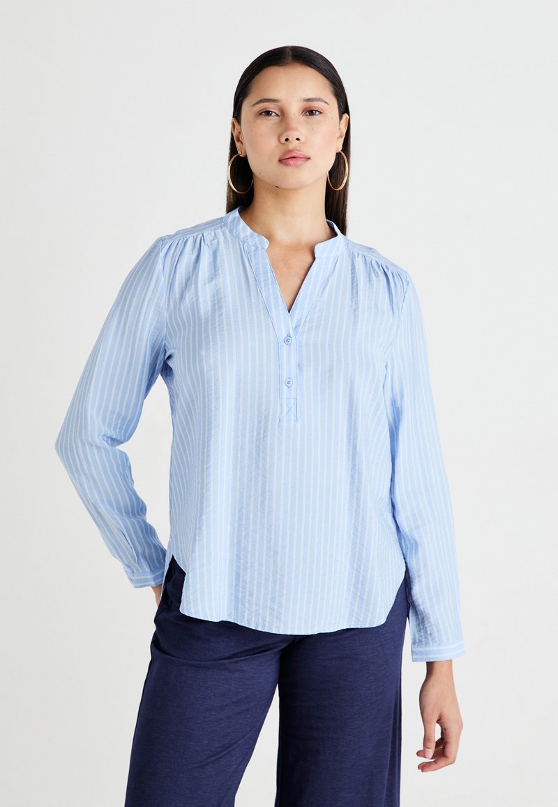 Camisa de mangas largas azul claro con rayas blancas verticales, una tapa de botones parcial y un dobladillo redondeado con ligero fruncido en los hombros.