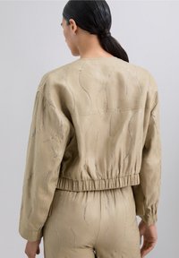 Chaqueta de un beige claro con cinturilla elástica, adornos curvados de plata, mangas largas y un diseño de corte relajado.