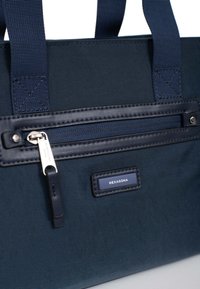 Borsa tote in tessuto blu navy con superficie testurizzata, dotata di tasca con zip, etichetta logo embossata e manici intrecciati resistenti.