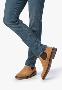 Jambes croisées portant un jean bleu et des bottines Chelsea en daim beige avec des panneaux élastiques noirs sur les côtés, sur fond blanc.