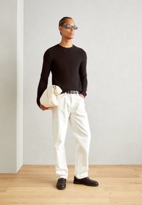 Svart ribbet langermet skjorte, hvite løstsittende bukser, brune loafers med kjededetalj, holder en beige veske, minimalistisk bakgrunn.