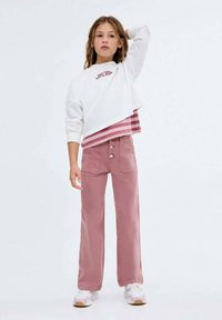 Bela skrajšana dukserica z rdečim napisom in prugastim robom, v kombinaciji z visokimi rožnatimi širokimi hlačami z sprednjimi žepi in gumbom.