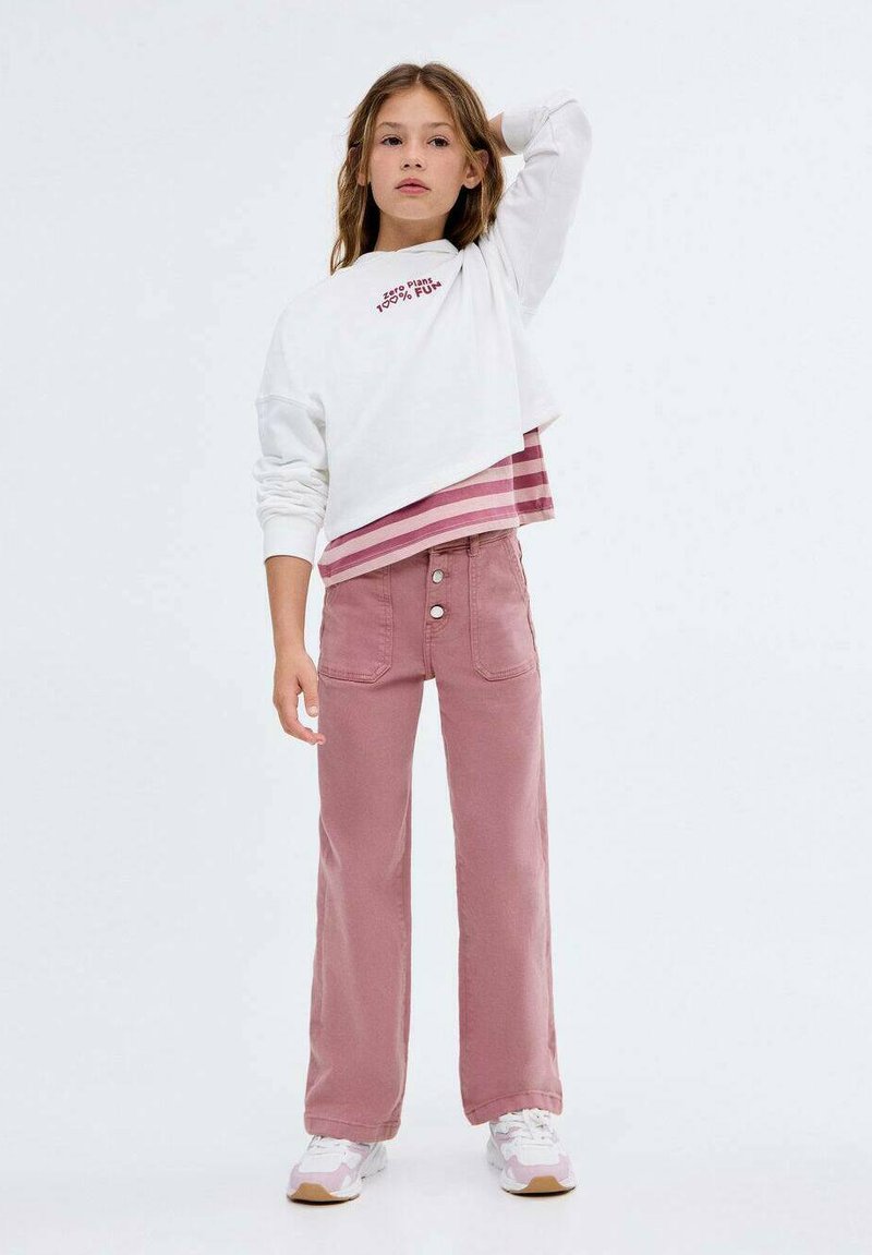 Bela skrajšana dukserica z rdečim napisom in prugastim robom, v kombinaciji z visokimi rožnatimi širokimi hlačami z sprednjimi žepi in gumbom.