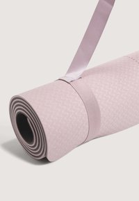 Joga prostirka svijetlo ružičaste boje s teksturiranom površinom. Rolana s tamno sivom donjom stranom, osigurana odgovarajućom trakicom.