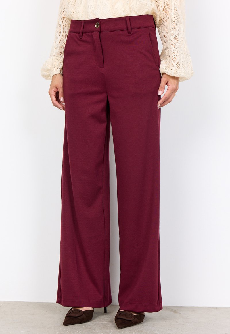 Frau in burgunderfarbenen, hochtaillierten, weit geschnittenen Hosen, kombiniert mit einer cremefarbenen, langen Spitzenbluse mit langen Ärmeln und braunen, spitzen Absätzen.