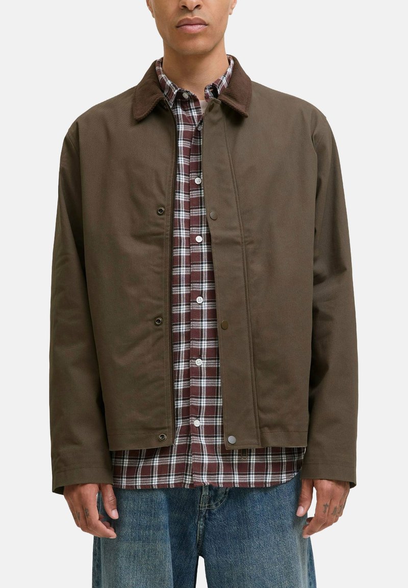 Chaqueta de lona marrón con cuello de pana, cierre de botones, superpuesta sobre una camisa de cuadros roja y blanca, combinada con vaqueros azules.