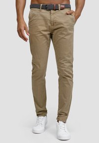 Chinos caqui de corte ajustado, que incluyen un cinturón negro tejido, bolsillos laterales y una textura suave. Combinados con zapatillas blancas.