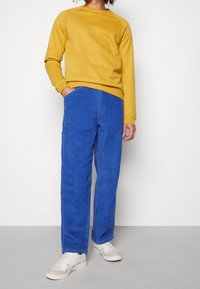 Gul sweatshirt med rund halsringning, i kombination med blå kordflortbyxor och vita sneakers. Byxorna har en fin ribbad textur och sidofickor.