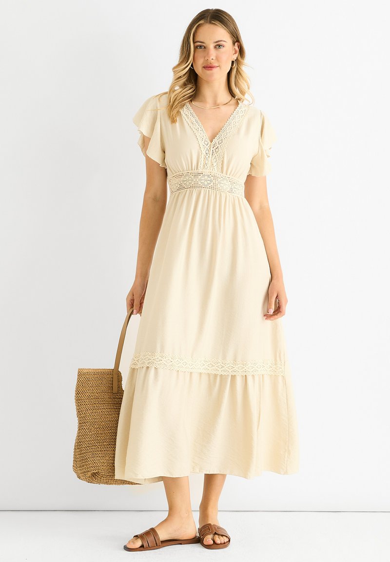 Beige, flowy maxi dress with short ruffle sleeves, lace detailing at the neckline and waistband, and a tiered hem. Worn with brown sandals.