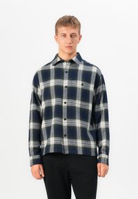 Young man facing forward wearing a black and white plaid button-up shirt and black pants against a plain white background.