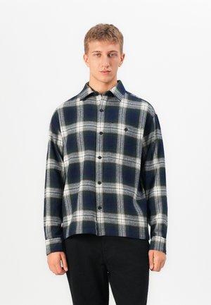 Young man facing forward wearing a black and white plaid button-up shirt and black pants against a plain white background.