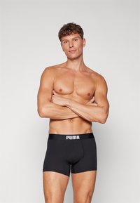 Black boxer shorts featuring a wide elastic waistband with "PUMA" logo. Smooth texture, fitted design, and knee-length cut.