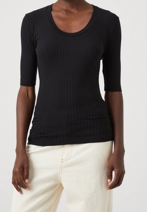 Person trägt ein eng anliegendes, schwarzes, geripptes Kurzarmshirt mit rundem Ausschnitt und offwhitefarbene, hoch taillierte Hose, steht vor einem schlichten Hintergrund.