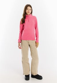 Roze fleece half-zip pullover met hoge kraag, gecombineerd met beige sneeuwbroeken en zwarte geïsoleerde laarzen, op een witte achtergrond.