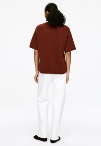 Rostfarbener kurzärmeliger Pullover aus weichem, strukturiertem Material, kombiniert mit weißen gerade geschnittenen Hosen und schwarzen Slippern.