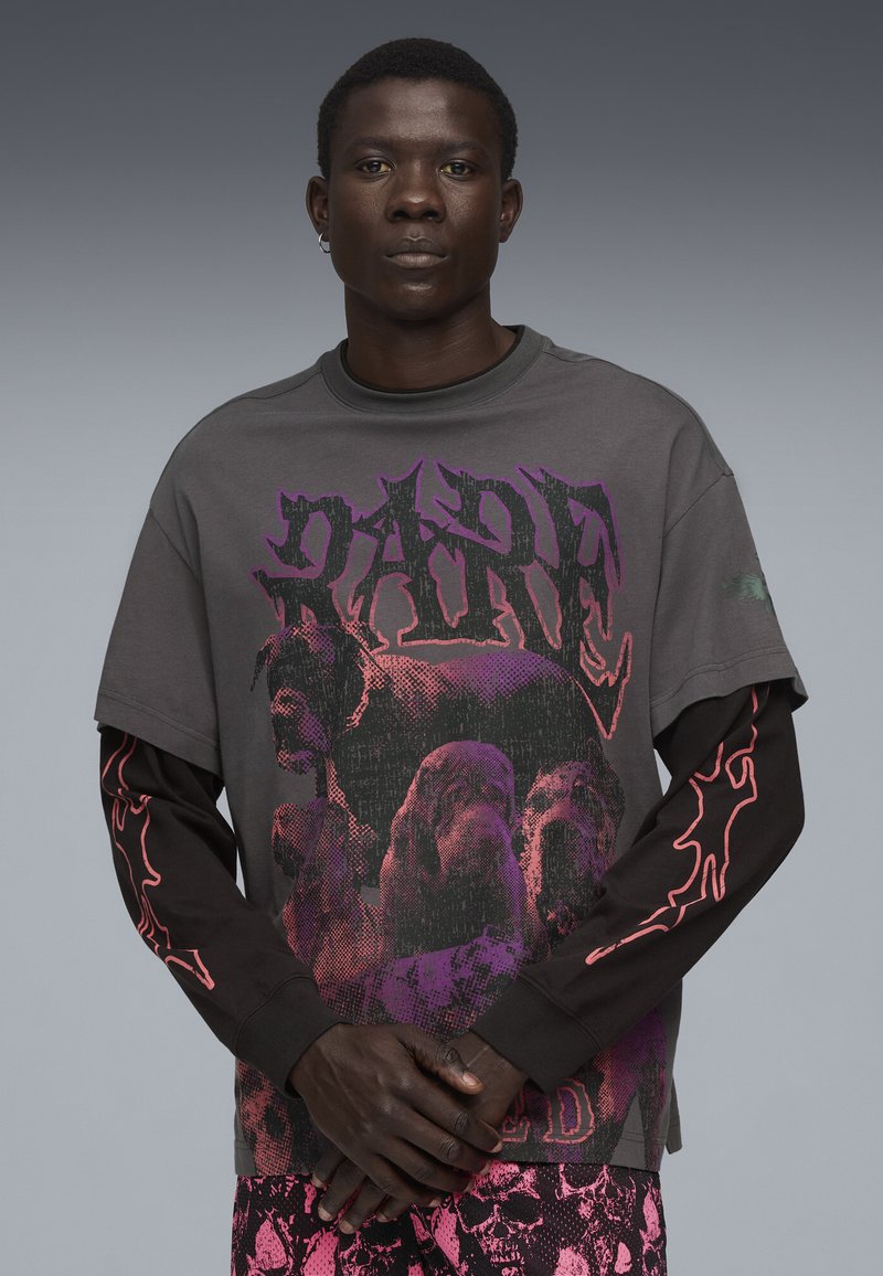 Grafika w oversize'owym t-shircie w ciemnoszarym kolorze, z kolorowymi wzorami psów i odważnym napisem. Długie czarne rękawy z czerwonymi akcentami w kształcie płomieni.