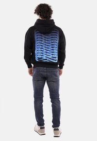 Felpa nera con un motivo a onda in gradiente blu sulla schiena, abbinata a jeans blu e sneakers chiare.