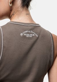 Bruine geribbelde mouwloze top met lichtgrijze stiksel en geborduurde tekst op de achterkant die luidt: "GOOD FOR NOTHING BRITISH ATELIER EST 2011."
