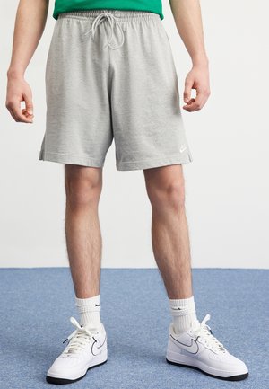Osoba nosząca jasnoszare spodenki Nike, białe buty sportowe Nike z czarnymi podeszwami oraz białe skarpetki stojąca na niebieskim dywanie.