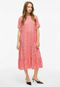 Robe maxi rose à motifs floraux, avec des manches courtes et une jupe à volants ; fabriquée en tissu léger ; modèle portant des mules à talons noirs.