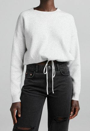 Personne portant un pull en maille gris clair avec taille à cordon de serrage et jean noir taille haute déchiré, debout devant un fond gris uni.